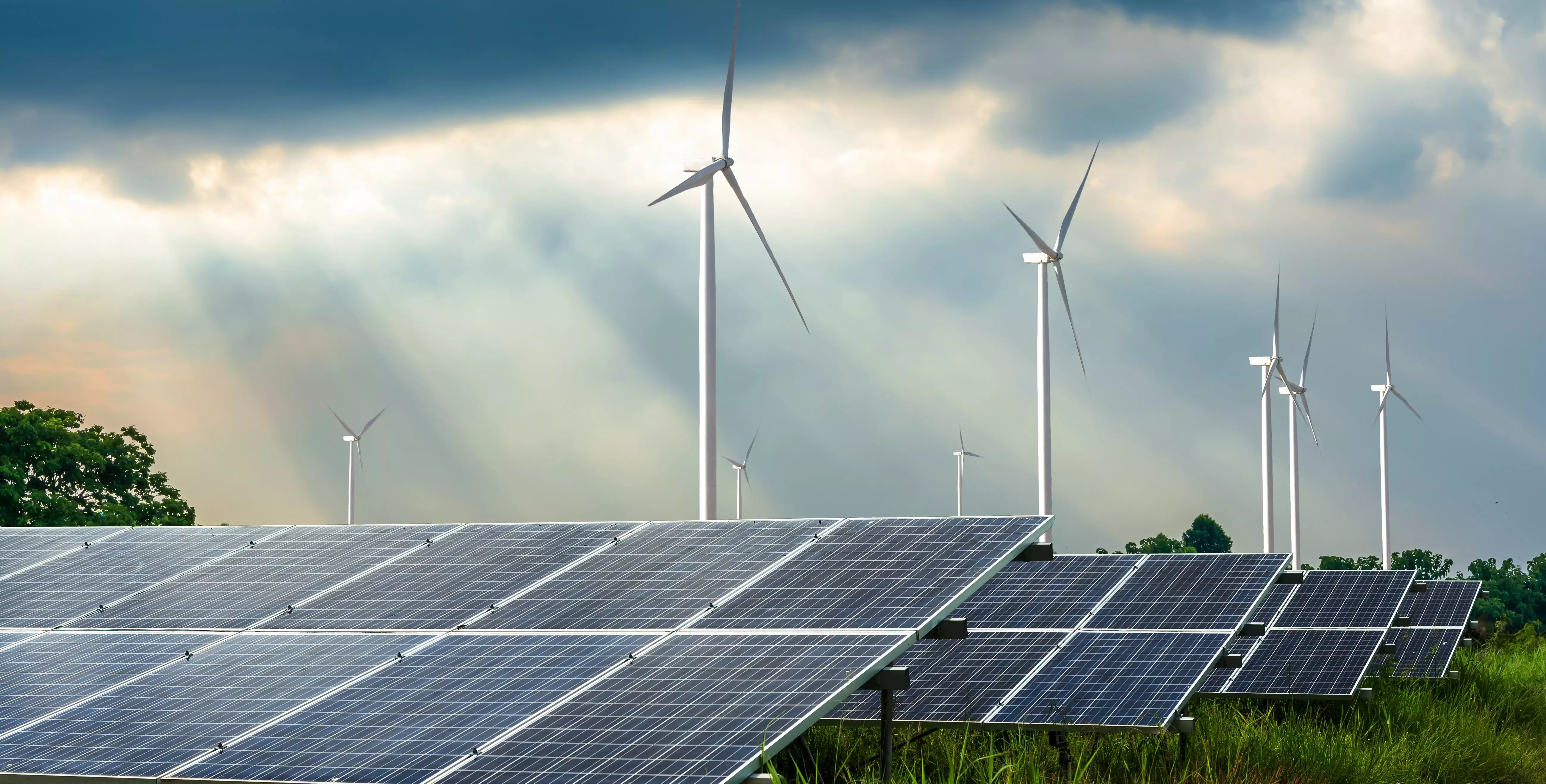 PV-Anlagen vor Windrädern auf Wiese vor bewölktem Himmel mit Sonnenstrahlen und Bäumen im Hintergrund